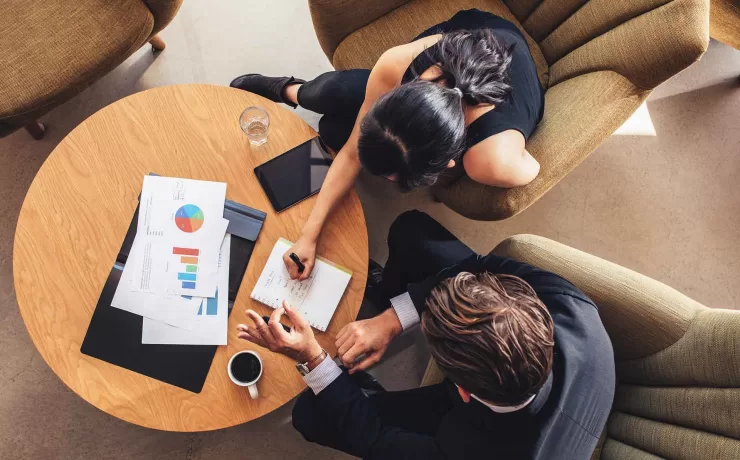 Due persone confrontano documenti di lavoro durante un incontro, per individuare le esigenze del cliente